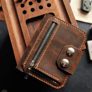 May include: A brown leather wallet with a zippered pocket and two silver metal snaps. The wallet has a distressed look and is made of high-quality leather.