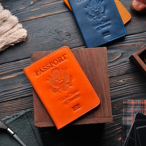 May include: Two leather passport covers, one orange and one blue, with the words "PASSPORT" and "United States of America" embossed on the front. The covers are lying on a wooden surface.