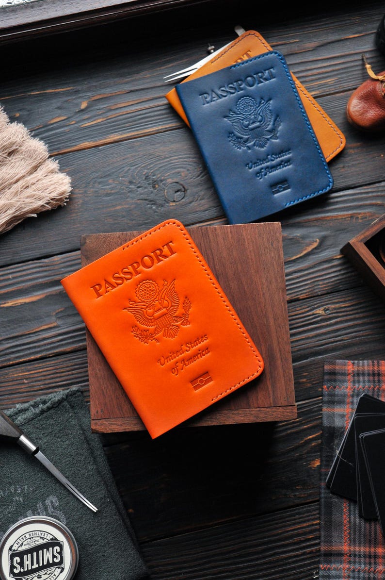 May include: Two leather passport covers, one orange and one blue, with the words "PASSPORT" and "United States of America" embossed on the front. The covers are lying on a wooden surface.