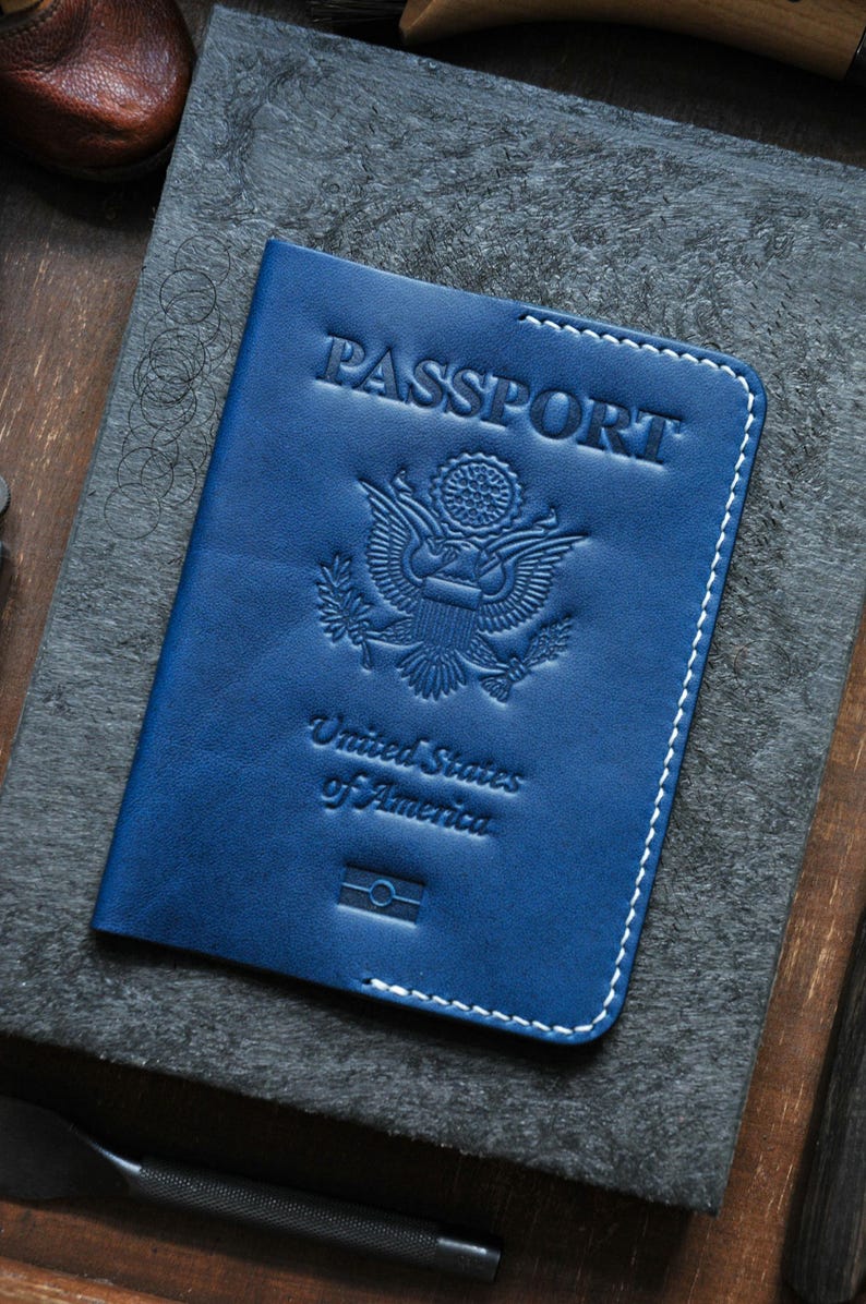 May include: A blue leather passport cover with white stitching. The cover has the word "PASSPORT" embossed on the front, along with the United States seal and the words "United States of America".