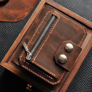 May include: A brown leather wallet with a zippered pocket and two silver snaps. The wallet is displayed in a wooden box.