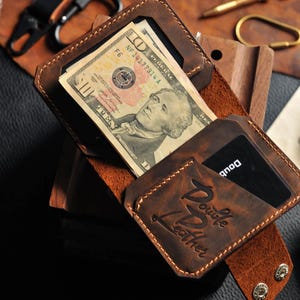 May include: A brown leather wallet with a flap closure and a card slot. The wallet has a stitched design and the text "Double Leather" embossed on the front. The wallet is open and contains a ten dollar bill and a credit card.