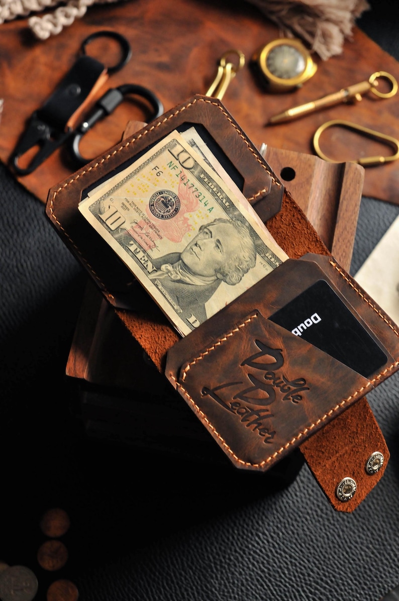 May include: A brown leather wallet with a flap closure and a card slot. The wallet has a stitched design and the text "Double Leather" embossed on the front. The wallet is open and contains a ten dollar bill and a credit card.