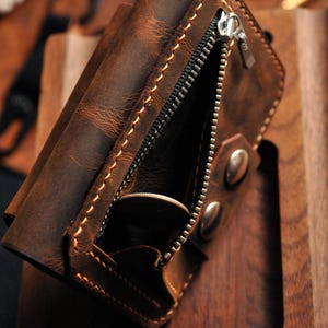 May include: A brown leather wallet with a zippered pocket and two snap closures. The wallet is made of distressed leather and has a rustic look.