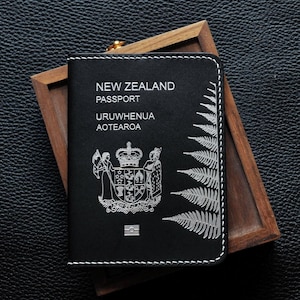 Może przedstawiać: Czarny pokrowiec na paszport z białymi przeszyciami, z napisem "NEW ZEALAND PASSPORT" i "URUWWHENUA AOTEAROA". Posiada srebrny wzór paproci i herb. Paszport spoczywa w drewnianej tacy.