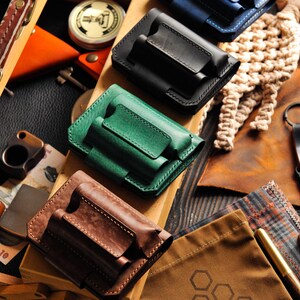 May include: Five leather cases in different colors, including blue, black, green, and brown, are arranged on a wooden surface. Each case has two slots for holding small items, such as knives or tools.