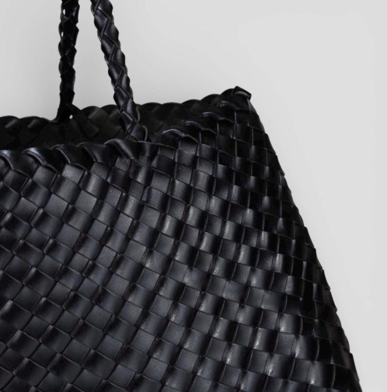May include: Black woven tote bag with braided handles. The bag features a basket weave design, with a structured shape. The handles are also braided. The bag is made of a durable material.