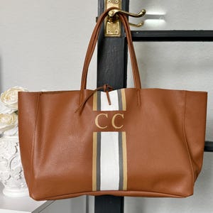 May include: A brown tote bag with a vertical stripe design in white, gold, and grey. The bag features the gold initials "CC" and has long brown handles. The bag is hanging on a black door frame with a gold handle.