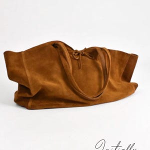 Puede incluir: Un bolso tote grande y abierto de ante marrón cálido. El bolso tiene dos asas largas y finas y un cierre de cordón en la parte superior. El bolso está sobre una superficie blanca. La marca "Initially Leather" está escrita en cursiva en la parte inferior.