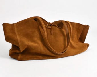 May include: A large, open-top tote bag in a warm brown suede. The bag has two long, thin handles and a drawstring closure at the top. The bag is sitting on a white surface. The brand name "Initially Leather" is written in cursive at the bottom.