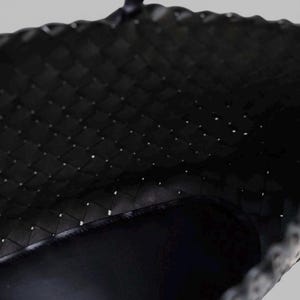 May include: Close-up of a black woven tote bag. The bag's interior is visible, showcasing a dark, smooth lining. The woven pattern creates a textured surface, with small gaps between the woven material.