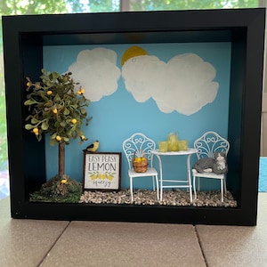 Könnte beinhalten: Ein Miniatur-Diorama mit schwarzem Rahmen, das einen Zitronenbaum, einen weißen Tisch und Stühle, eine Katze und ein Schild mit der Aufschrift "Easy Peasy Lemon Squeezy" zeigt.