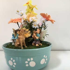 Może przedstawiać: Jasnoniebieska ceramiczna doniczka z białymi odciskami łapek wokół krawędzi. Doniczka jest wypełniona zielonym mchem, sztucznymi kwiatami i dwiema figurkami wróżek. Jedna wróżka trzyma małą figurkę psa, a druga jedzie na większej figurce psa.