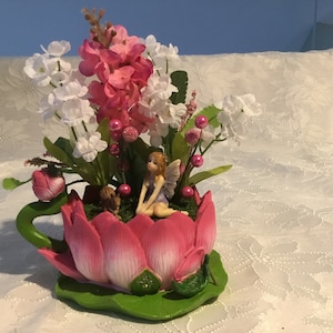 May include: A pink and white flower arrangement in a pink lotus-shaped planter with two fairies sitting on the rim. The planter is on a white tablecloth.
