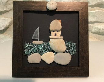 May include: A framed artwork made of small stones and glass depicting a house and a sailboat on a beach. The house is made of stacked stones and has a small window and a door. The sailboat is made of a single piece of glass. The beach is made of small stones and glass.