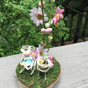 Könnte beinhalten: Eine Miniatur-Szene mit einer weißen Hasenfigur, die in einem weißen Schaukelstuhl sitzt, einer gelben Kükenfigur in einem geflochtenen Korb und einem Blumenarrangement mit rosa und weißen Blumen auf einem Holzsockel.
