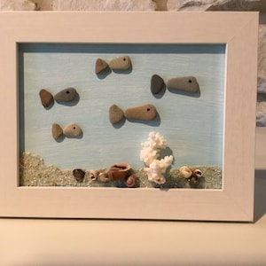 May include: A framed artwork featuring a blue background with white sand and seashells at the bottom. Five fish made of stones are arranged in a swimming pattern. A white coral piece is in the center.