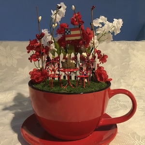 Puede incluir: Una taza de té roja con un platillo, llena de una escena de jardín en miniatura. El jardín incluye una valla blanca de piquetes, dos sillas rojas, una mesa, una cesta y flores rojas y blancas.