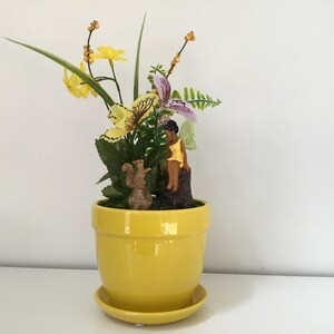 Może przedstawiać: Żółta ceramiczna doniczka z podstawką, przedstawiająca małą figurkę wróżki siedzącą na pniu drzewa, figurkę wiewiórki i kompozycję kwiatową z żółtymi i fioletowymi kwiatami.