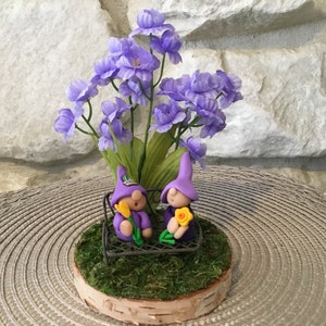 May include: Two purple gnomes sitting on a metal bench with yellow flowers in their hands. The gnomes are surrounded by purple flowers and green foliage on a birch wood base.