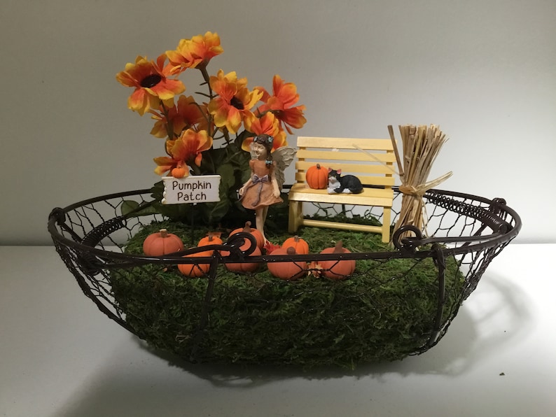 Peut inclure: Une sc&egrave;ne de jardin miniature de f&eacute;es dans un panier en fil de fer. La sc&egrave;ne comprend une figurine de f&eacute;e, un petit banc en bois, une figurine de chat noir, une pancarte indiquant "Pumpkin Patch", des citrouilles orange et un bouquet de bl&eacute; s&eacute;ch&eacute;.