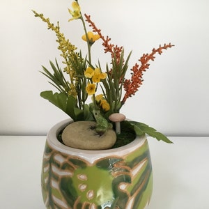May include: A ceramic pot with a green and white pattern, containing artificial flowers and foliage. A small frog ornament rests on a stone, beside a mushroom. The arrangement features yellow, orange, and green colours.