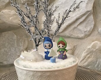 Può includere: Un vaso di ceramica bianca con un motivo a fiocco di neve, riempito di glitter bianco e una piccola scena innevata. Due piccole figurine fatte a mano di persone stanno nella neve, una con un cappello blu e l'altra con un cappello verde. Un piccolo albero bianco e scintillante si trova tra loro.