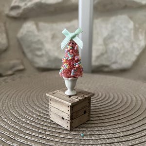 Op de afbeelding: Een miniatuur decoratieve boom met een roze basis en kleurrijke, kraalachtige ornamenten, bekroond met een lichtgroene strik. Hij staat in een witte vaas op een kleine, rustieke houten standaard. De standaard staat op een geweven, cirkelvormige mat.