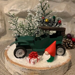 Könnte beinhalten: Ein grünes Oldtimer-Auto, das einen kleinen, beflockten Weihnachtsbaum mit roten und grünen Ornamenten transportiert. Ein Zwerg mit roter Mütze sitzt neben einem verpackten Geschenk mit einer roten Schleife auf einer Birkenholzscheibe.