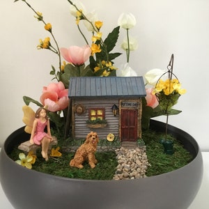 Op de afbeelding: Een miniatuur feeentuin scène in een grijze pot. Een klein houten huis met een blauw dak en de woorden "Potting Shed" aan de zijkant. Een fee figuur zit op een bankje, een klein hondje figuur zit in het gras en er zijn gele en roze bloemen.