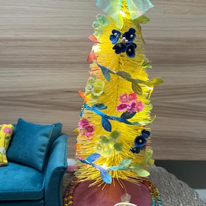 Puede incluir: Un caprichoso árbol decorativo amarillo adornado con flores coloridas, detalles de hojas azules y un lazo azul claro. El árbol está decorado con pequeñas luces y se asienta sobre una base rosa. Una pequeña cesta de caramelos en la base.