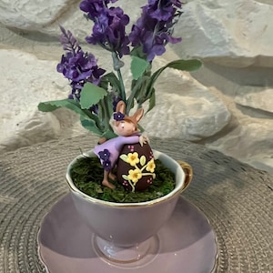 Puede incluir: Una pequeña figurita de cerámica de un conejito sosteniendo un huevo de Pascua de chocolate decorado con flores amarillas, sentada en una taza de té morada con un platillo. La taza de té y el platillo están decorados con un borde dorado. El conejito lleva un vestido morado con una flor rosa en la cabeza. La taza de té está rodeada de flores de lavanda moradas.