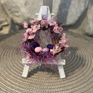 Pode incluir: Uma guirlanda de Páscoa em miniatura com flores cor-de-rosa, ovos roxos e cor-de-rosa e um ninho brilhante rosa e roxo, exibida em um cavalete branco.