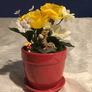 Puede incluir: Una maceta de cerámica roja con un arreglo de flores blancas y amarillas. Una pequeña figura de ardilla está sentada en una roca en la maceta, sosteniendo una nuez. También hay un pequeño hongo rojo y blanco en la maceta.