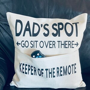 May include: A white throw pillow with black text that reads "Dad's Spot - Go Sit Over There - Keeper of the Remote". The pillow has a pocket on the front that holds a black remote control.