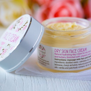 Peut inclure: Un pot blanc avec une étiquette florale rose et dorée qui indique "DRY SKIN FACE CREAM". Le pot contient une crème jaune et est ouvert, révélant la crème à l'intérieur. L'étiquette liste les ingrédients, dont de l'eau distillée, de la calendula, de la cire d'abeille et des huiles de vanille et de patchouli.