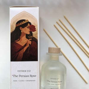 May include: A reed diffuser set featuring a white rectangular box and a frosted glass bottle with a wooden stopper. The box displays an illustration of a woman with a red rose and the text Esther 2:12 The Persian Rose. Five wooden reeds are included.