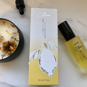 Könnte beinhalten: Ein Hautpflege-Produktset mit einer gelb getönten Serumflasche und einer passenden Schachtel, beide mit der Aufschrift "Citron Night Serum". Die Schachtel hat eine Zitronenillustration. Eine schwarze Kerze mit getrockneten Blumen ist ebenfalls vorhanden. Die Serumflasche ist 30 ml / 1 fl oz.