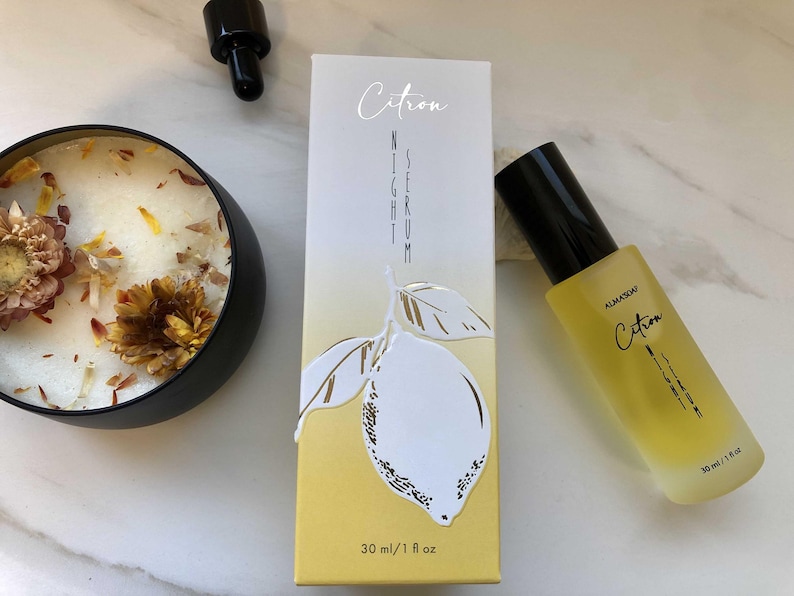 May include: A skincare product set featuring a yellow-tinted serum bottle and a matching box, both labeled "Citron Night Serum." The box has a lemon illustration. A black candle with dried flowers is also present. The serum bottle is 30 ml / 1 fl oz.