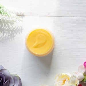 Pode incluir: Um pequeno pote redondo de creme amarelo sobre uma superfície de madeira branca. O pote é cercado por flores brancas e rosas.