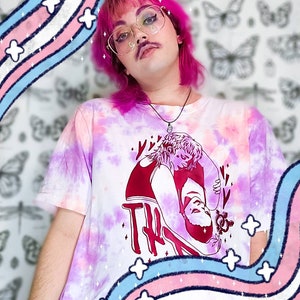 May include: A person with pink hair and a mustache is wearing a white tie-dye t-shirt with a red graphic of two people embracing. The graphic includes the text "Try" and a transgender symbol. The background is a white wall with a pattern of butterflies and dragonflies. The t-shirt is decorated with a rainbow-colored design.
