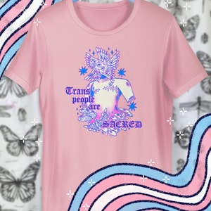 May include: A pink t-shirt with a graphic that reads "Trans people are sacred." The graphic features a stylized figure with a bird-like head and floral elements in blue and purple. The shirt is hanging on a blue hanger.