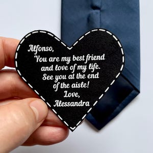 May include: A black heart-shaped tag with white stitching and text. The text reads: "Alfonso, You are my best friend and love of my life. See you at the end of the aisle! Love, Alessandra." A navy blue tie is in the background.