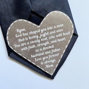 May include: A navy blue tie with a heart-shaped tag. The tag is light brown with white stitching and text that reads "Ryan, God has shaped you into a man...Love you forever & always, Mom."