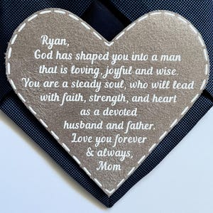 May include: A navy blue tie with a heart-shaped felt applique. The applique has a stitched border and contains white text that reads "Ryan, God has shaped you into a man...Love you forever & always, Mom."
