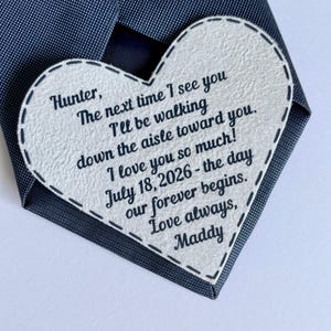May include: A dark blue textured necktie featuring a white heart-shaped patch stitched onto the fabric. The patch contains black printed text: Hunter, The next time I see you I'll be walking down the aisle toward you. I love you so much! July 18, 2026 - the day our forever begins. Love always, Maddy.