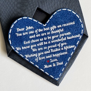 May include: A blue heart-shaped card with a white stitched border and a message written in white text. The message reads: "Dear John, You are one of the best gifts we received and we are so thankful God chose us to be your parents. We know you will be a wonderful husband. We are so proud of you. Wishing you and Kailee a lifetime of love and happiness. Love, Mom & Dad."