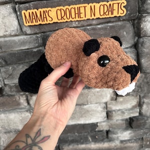 May include: A crocheted brown and black beaver plush toy with white teeth. The toy is being held by a hand with a tattoo.