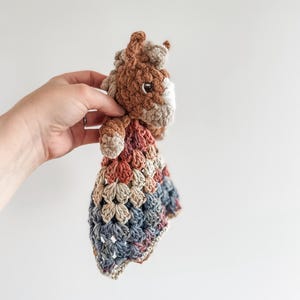 May include: A crocheted stuffed animal horse with a brown and beige body and a blue, beige, and brown blanket attached. The horse has a white face with black eyes.