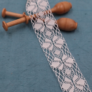 Peut inclure: Ruban de dentelle blanc avec un motif floral, présenté sur un fond bleu-vert. Deux bobines en bois sont visibles. La dentelle présente un motif ajouré, idéal pour les travaux de couture et d'artisanat.